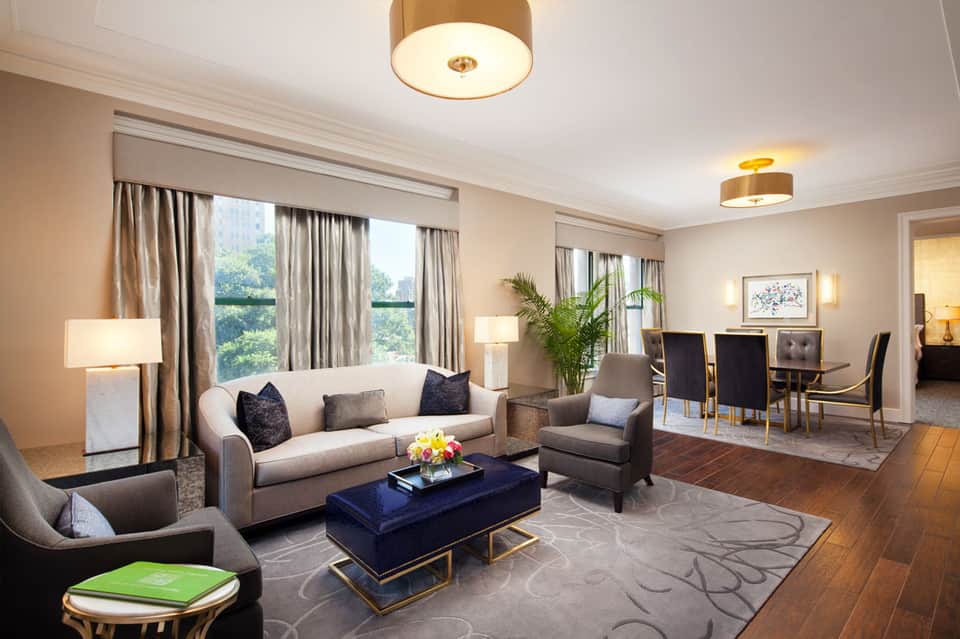 Luxury hotel suite living room with beige sofa, blue ottoman, floor-to-ceiling windows overlooking city skyline