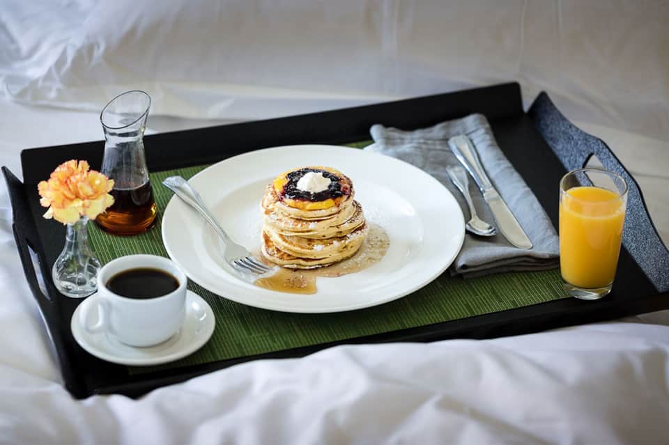 In-room breakfast tray with stacked pancakes, coffee, orange juice, and fresh flowers on a bed