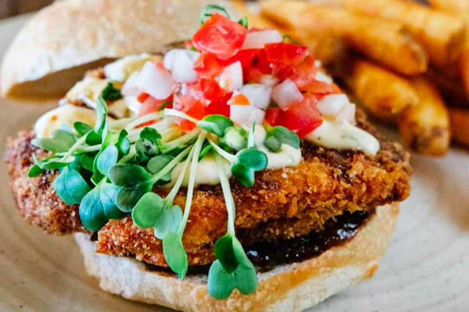 Gourmet chicken sandwich with microgreens, diced tomatoes, white sauce, and crispy coating on a bun