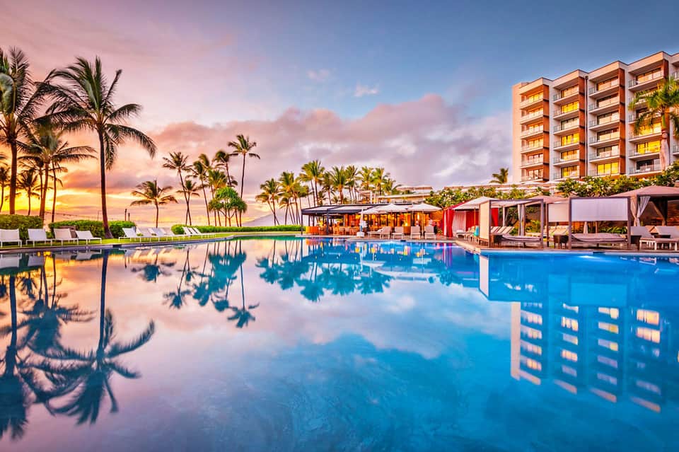 Resort pool at sunset with palm trees, colorful sky reflection, and multi-story hotel building in background