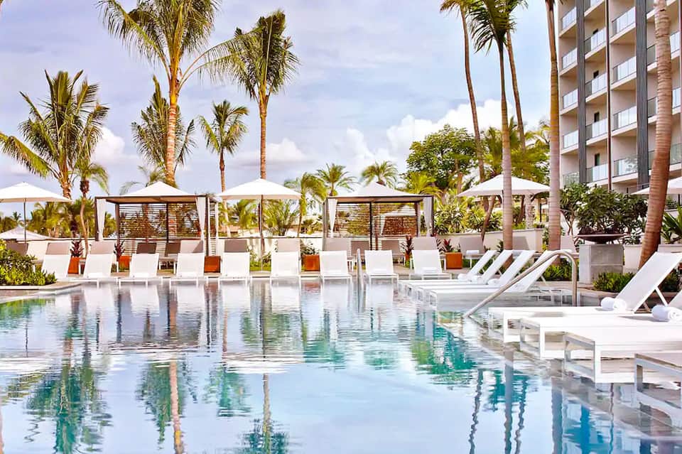 Luxury resort pool with white loungers, palm trees, and modern cabanas under blue sky