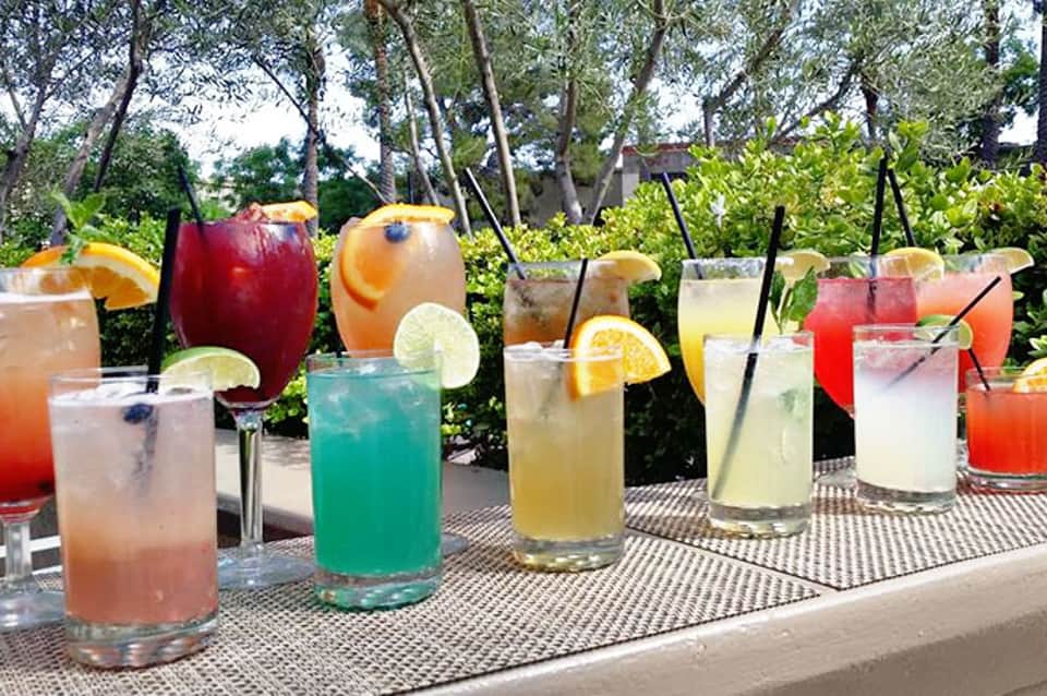Array of colorful cocktails with fruit garnishes displayed outdoors on a patterned table with trees in background