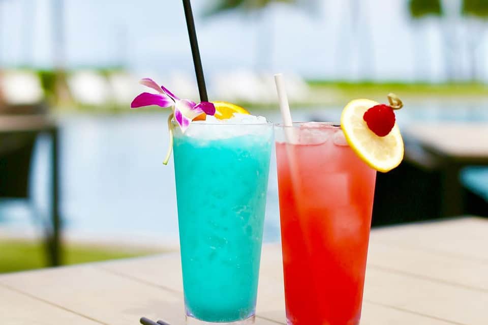 Two colorful tropical cocktails with garnish and straws on an outdoor deck near water