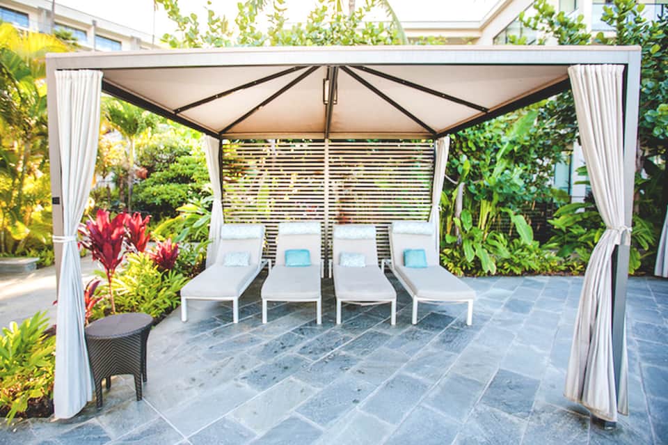 Elegant poolside cabana with white lounge chairs, curtains, and lush tropical landscaping