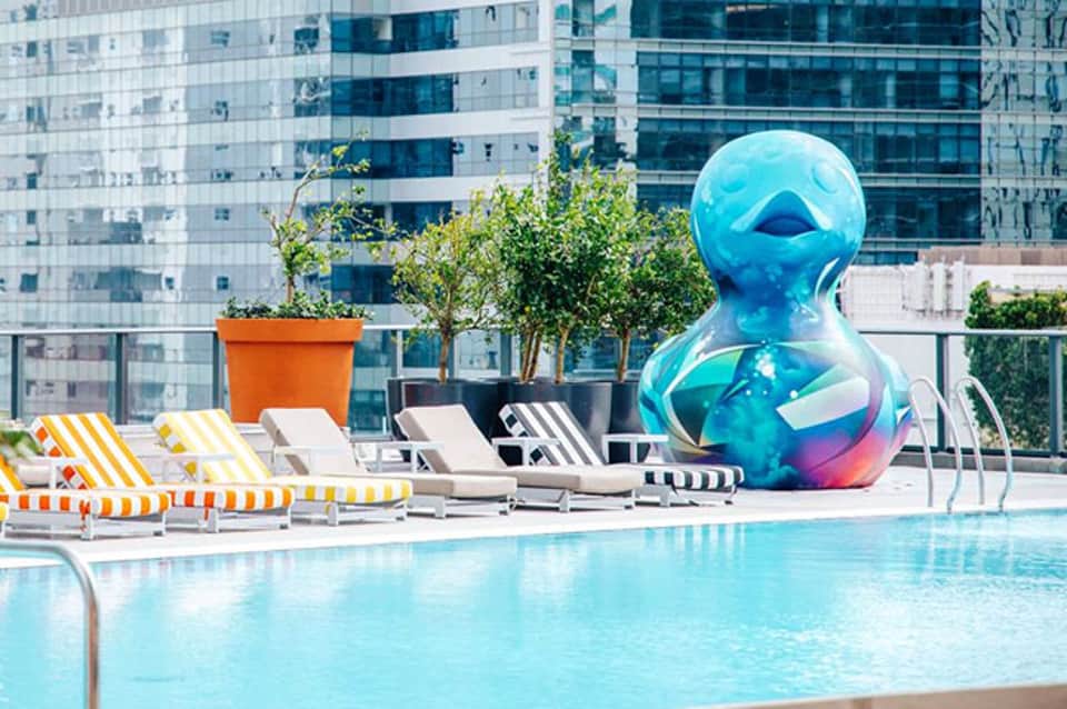 Modern rooftop pool with blue sculptural art, loungers, and downtown skyscrapers in background