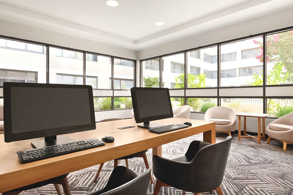 Modern office workspace with desktop computers, wooden desk, and large windows overlooking resort grounds