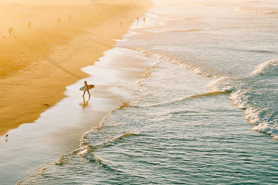 Surfer in wetsuit walking into waves on golden sandy beach at sunrise