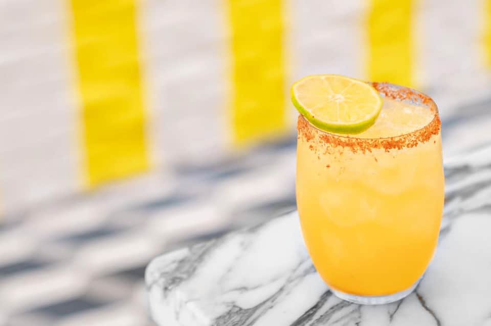 Yellow cocktail with lime slice and spiced rim on marble surface with striped umbrellas in background