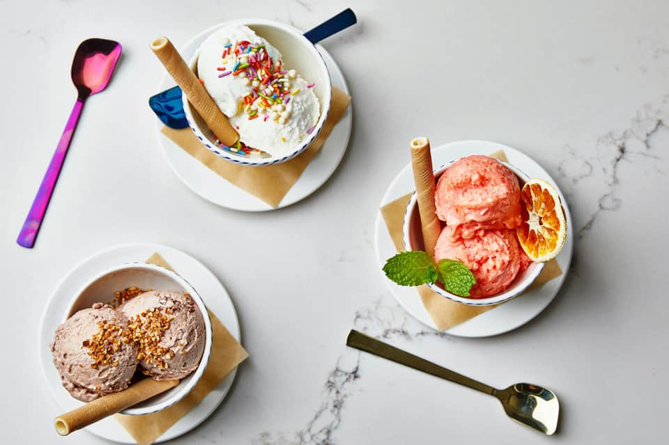 Three ice cream bowls with colorful sprinkles, chocolate, and grapefruit garnish on marble surface