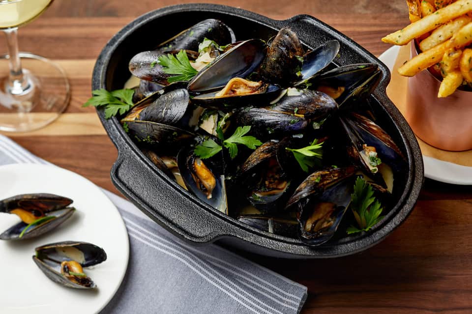 Black cast iron bowl of steamed mussels with parsley garnish, fries, and white wine on wooden table