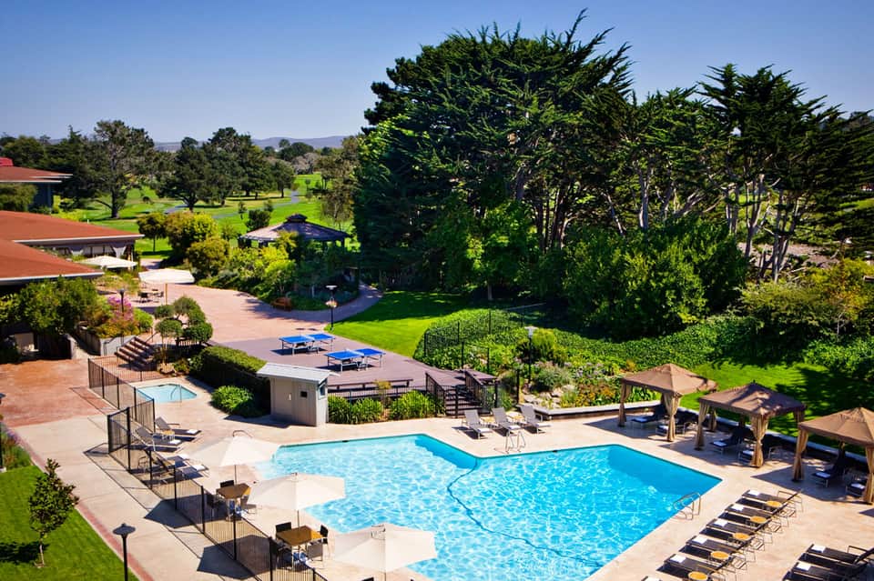 Aerial view of resort pool surrounded by manicured lawns, trees, and wooden structures with red-tiled roofs