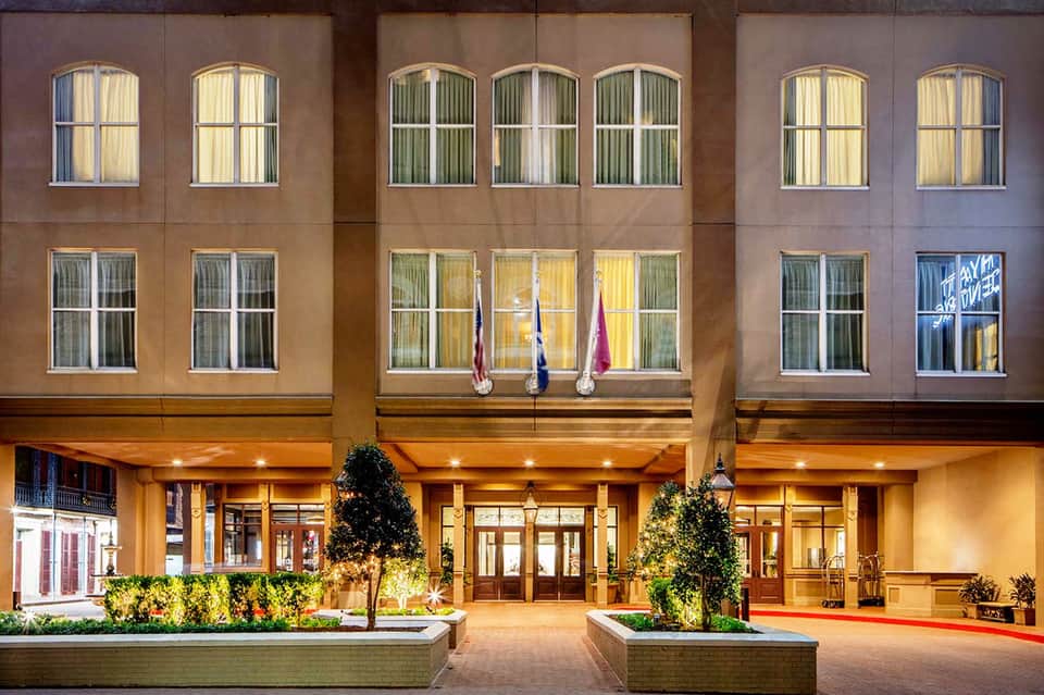 Hotel entrance at dusk with illuminated facade, large windows, and decorative planters flanking glass doors.