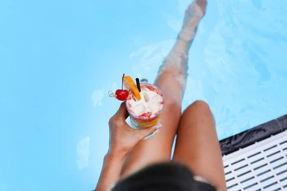 Person holding colorful cocktail with fruit garnish above pool