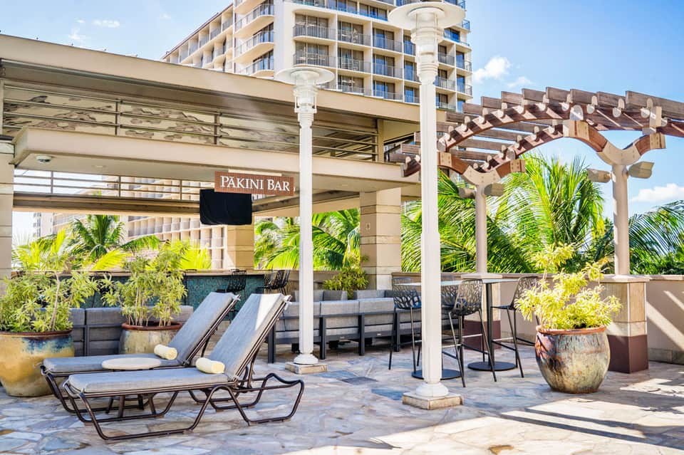 Outdoor Pakini Bar patio with lounge chairs, wooden pergola, potted plants, and high-rise hotel building