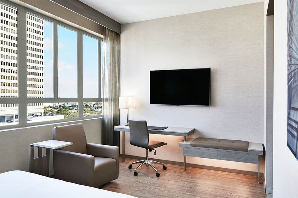 Modern hotel room with desk, office chair, flat-screen TV, armchair, and floor-to-ceiling windows overlooking city