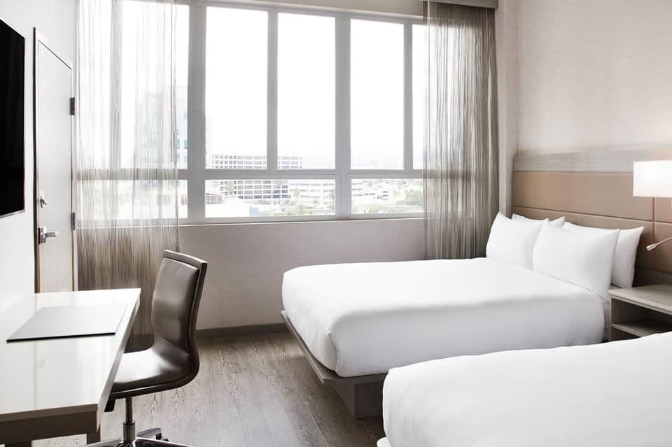 Modern hotel room with two beds, city skyline views through floor-to-ceiling windows, and contemporary furnishings