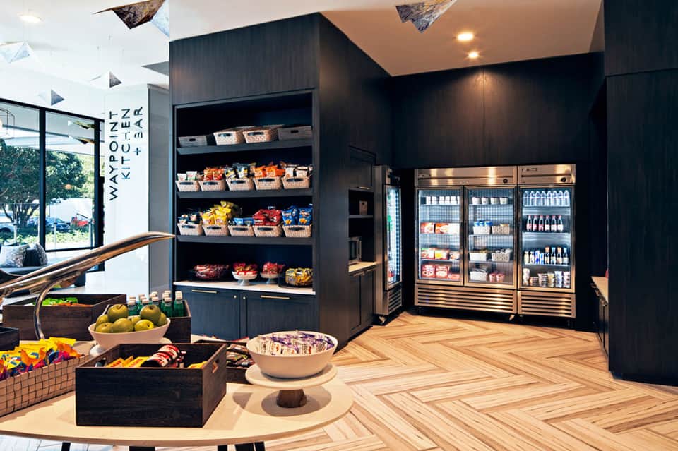 Modern resort marketplace with dark wood shelving, refrigerated beverage displays, fresh produce baskets, and herringbone wooden flooring