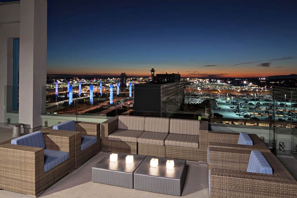 Rooftop lounge at dusk with wicker seating, illuminated blue columns, and cityscape views