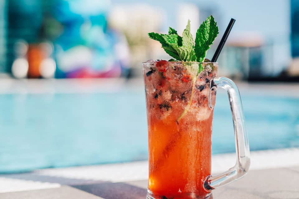 Red tropical cocktail with ice, mint, and black straw in glass mug by poolside