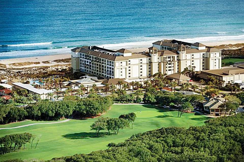 Beachfront resort with multi-story hotel buildings, manicured golf course, palm trees, and turquoise ocean waters.