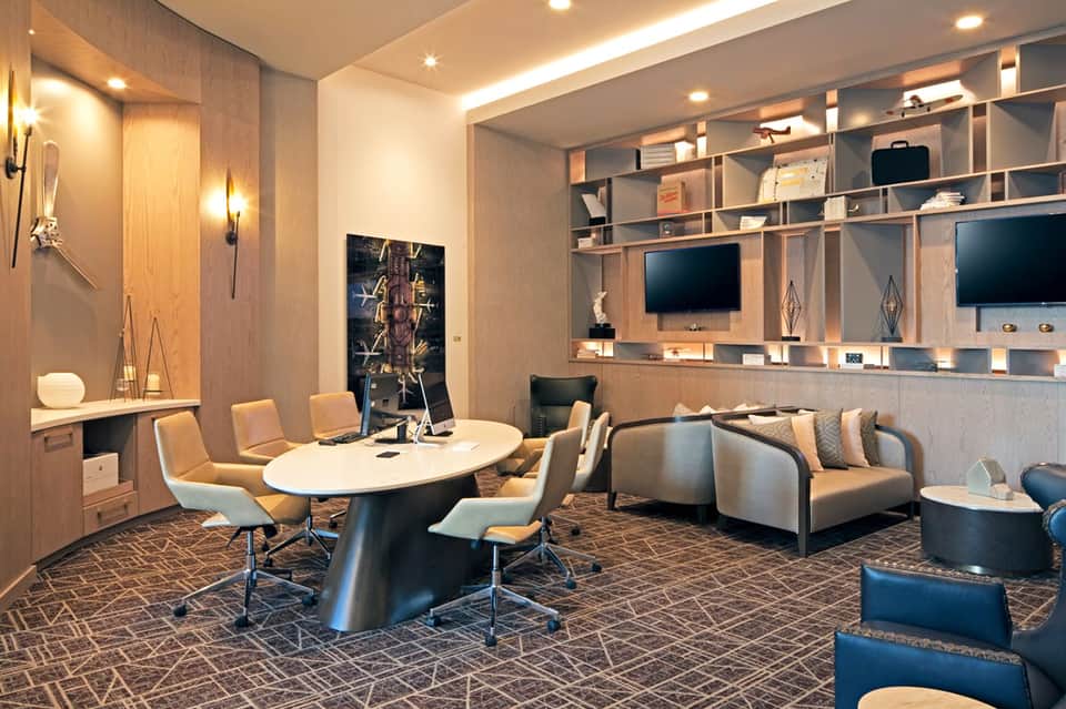 Modern hotel conference room with oval table, tan chairs, wood shelving, and dual monitors on wall