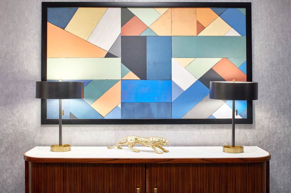 Modern geometric abstract artwork in blue, orange, and cream tones above wooden dresser with brass accents