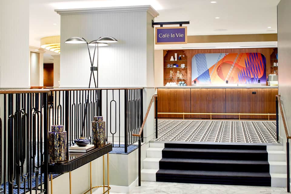 Modern hotel lobby with Café la Vie sign, black railing, decorative floor lamp, and colorful digital display above counter
