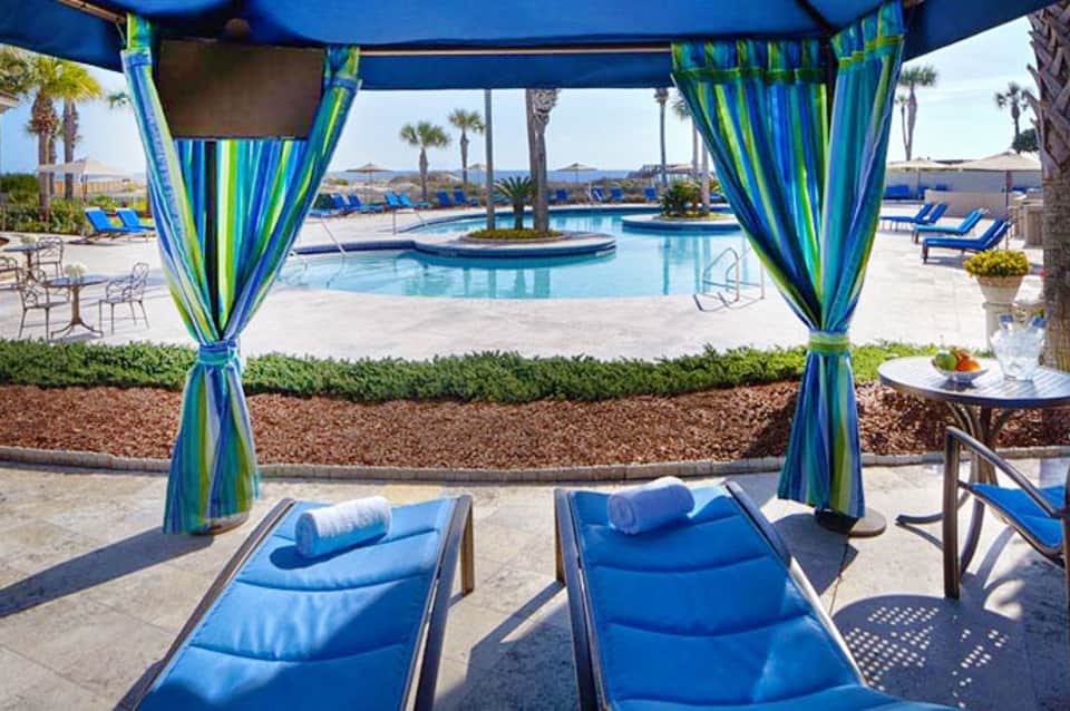 Beachside pool area with blue cabana and striped curtains overlooking the ocean with palm trees
