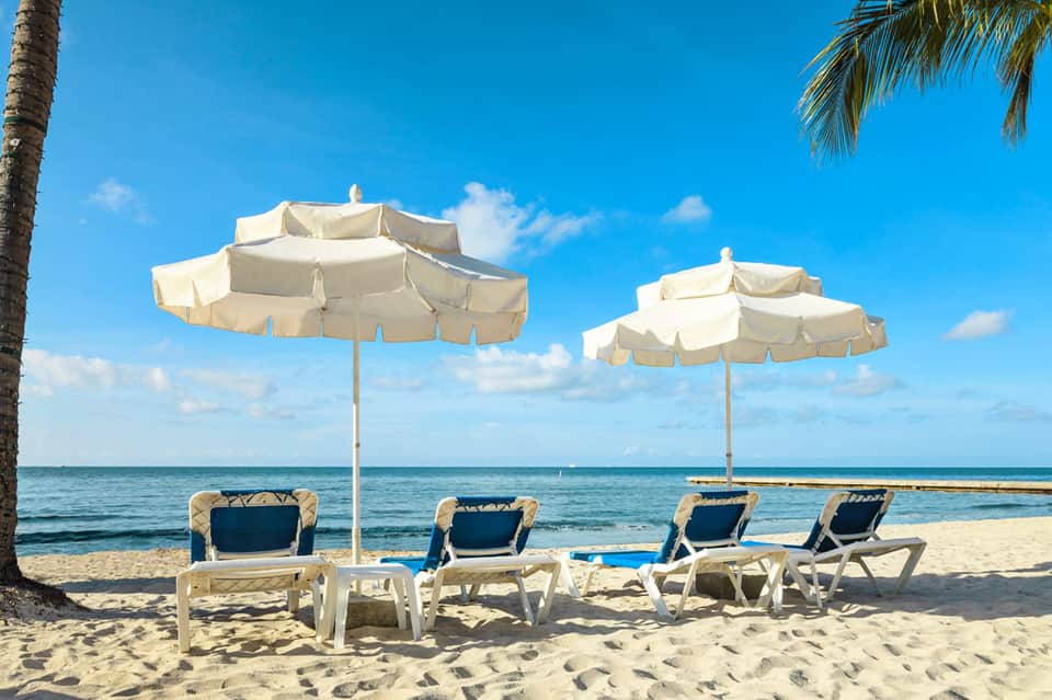 Beach lounge area with cream umbrellas, blue chairs, and palm trees overlooking turquoise ocean water