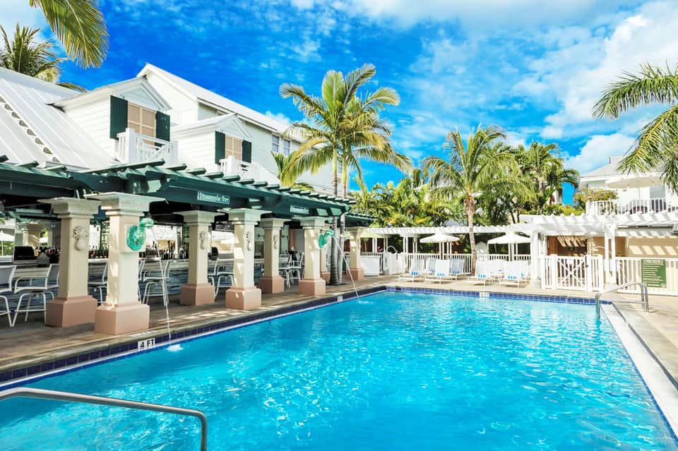 Bright blue resort pool with green pergola, white colonial-style main building, and palm trees under clear sky