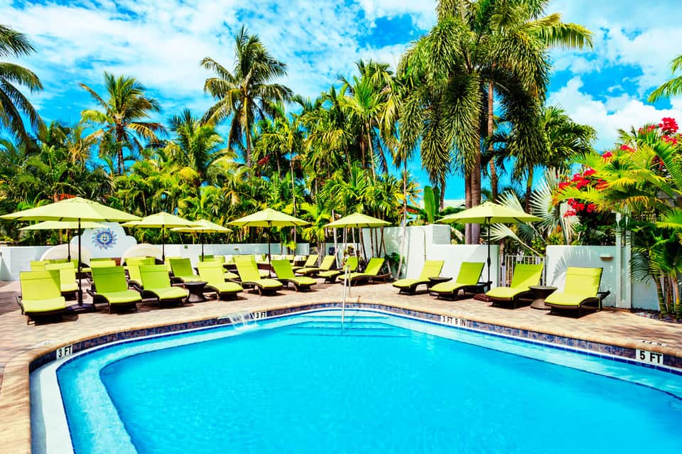 Tropical resort pool with lime green loungers, palm trees, and yellow umbrellas under blue sky