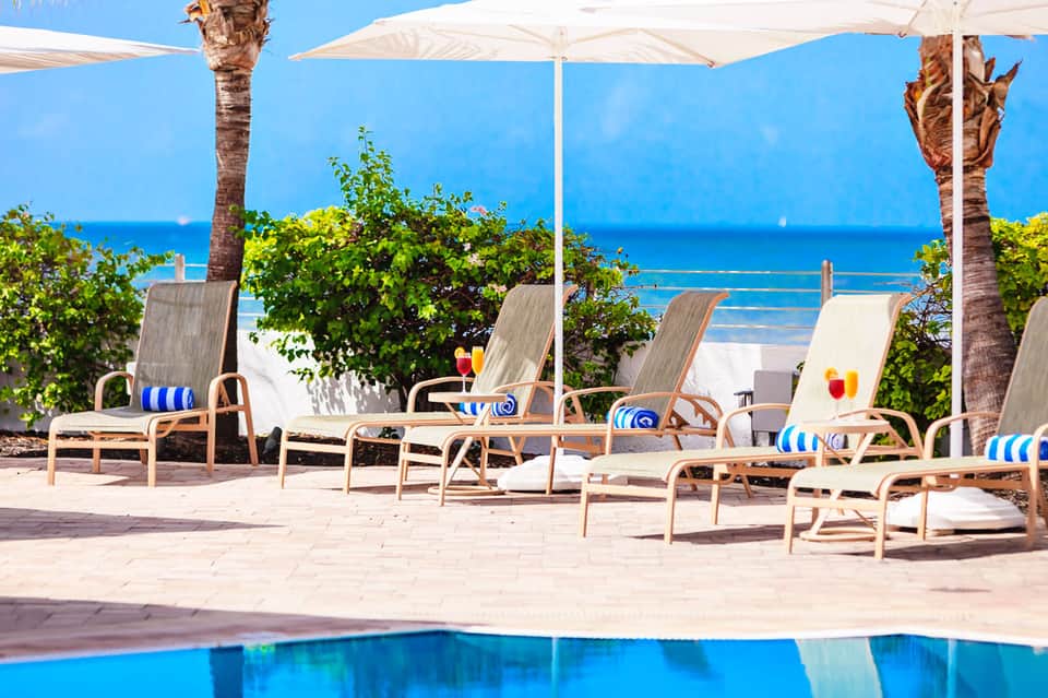 Beachfront lounge area with white umbrellas, palm trees, ocean view, and blue waters beyond