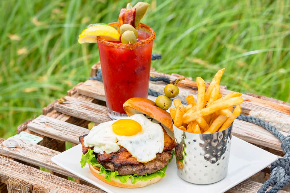 Gourmet burger with fried egg and bacon, fries in a cup, and red cocktail on rustic wooden table outdoors