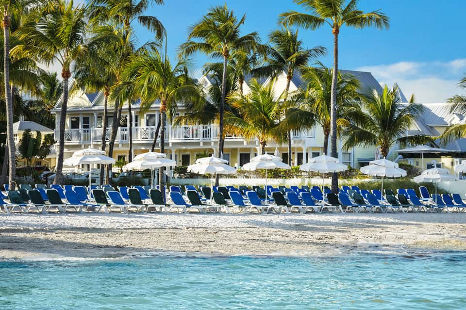 Beachfront resort with blue loungers, white umbrellas, palm trees, and colonial architecture