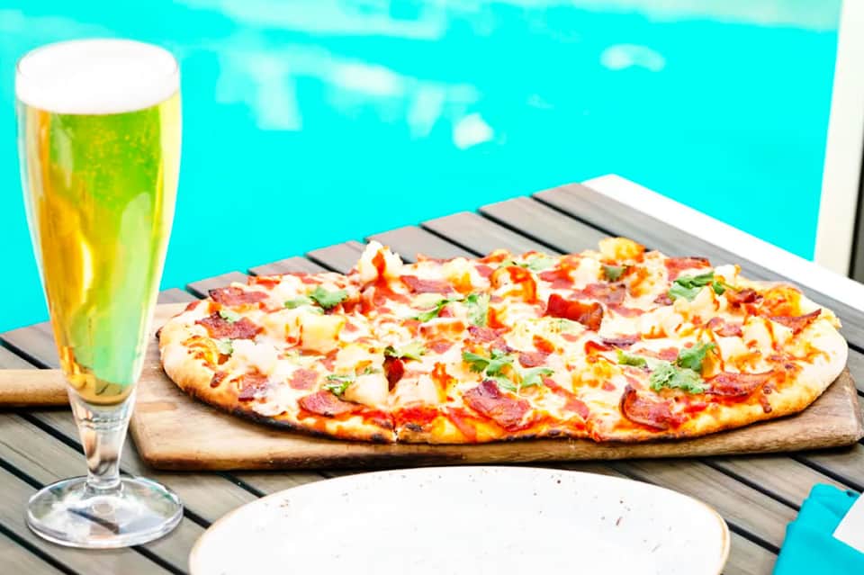 Pepperoni pizza on wooden board with beer glass overlooking turquoise pool water