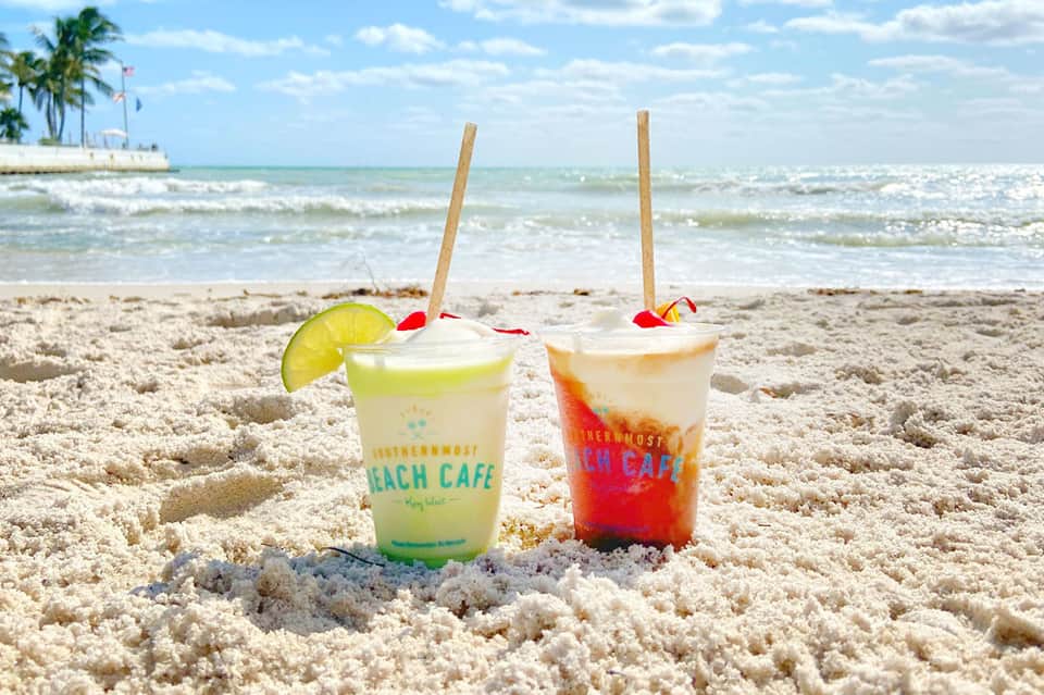 Two colorful beverages from Southernmost Beach Cafe sitting in sand with ocean and palm trees in background