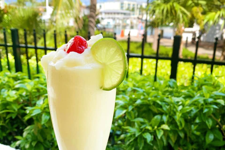 Cocktail with lime slice and cherry garnish on outdoor patio with palm trees and black fence