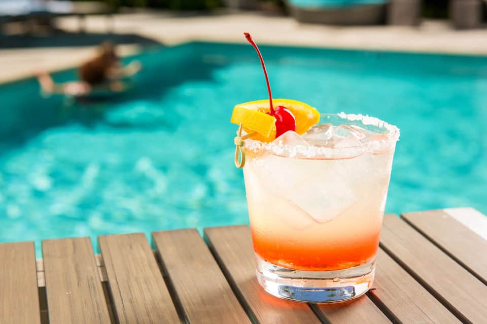 Colorful layered cocktail with cherry and pineapple garnish on poolside deck overlooking blue pool
