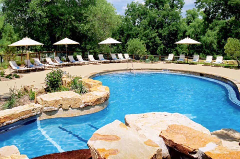 Curved resort pool with loungers, white umbrellas, and natural stone features surrounded by trees