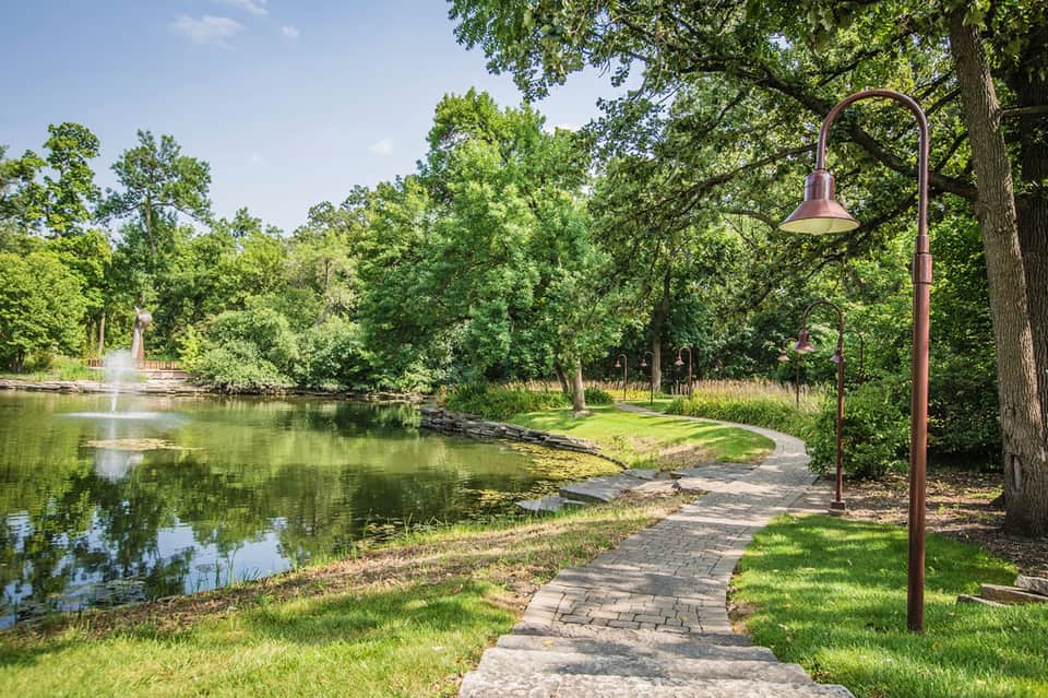 Scenic paved pathway along a calm pond surrounded by lush green trees and landscaping with a vintage street lamp