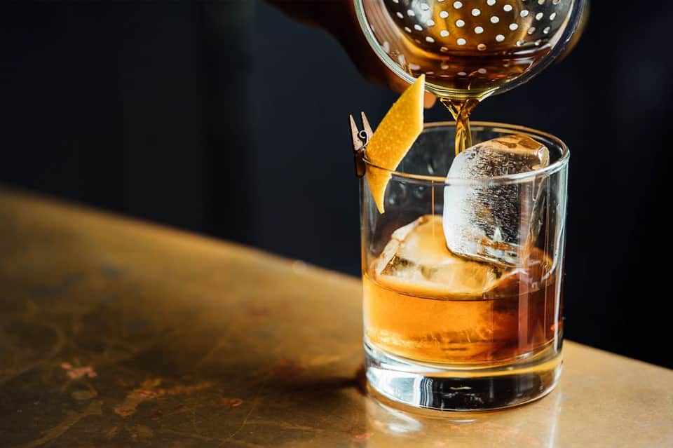 Whiskey being poured into a glass with ice and orange peel garnish on a dark surface