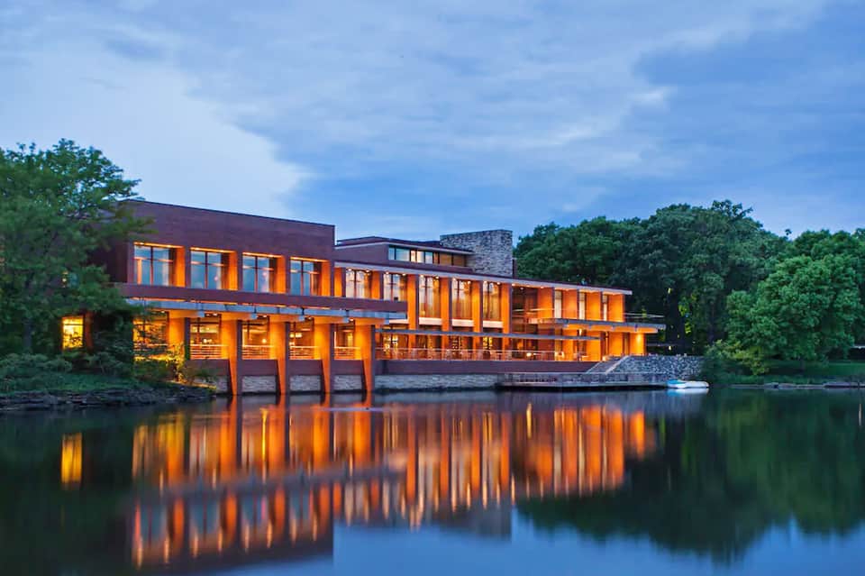 Modern waterfront building with orange-lit windows reflecting in calm river at dusk, surrounded by trees