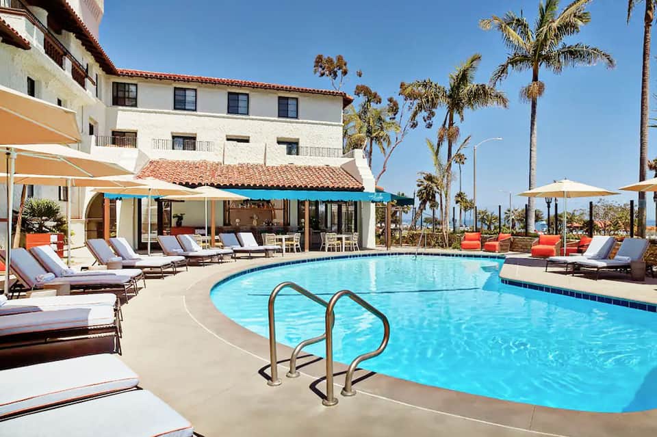 Resort pool area with lounge chairs, umbrellas, Spanish colonial building, and palm trees