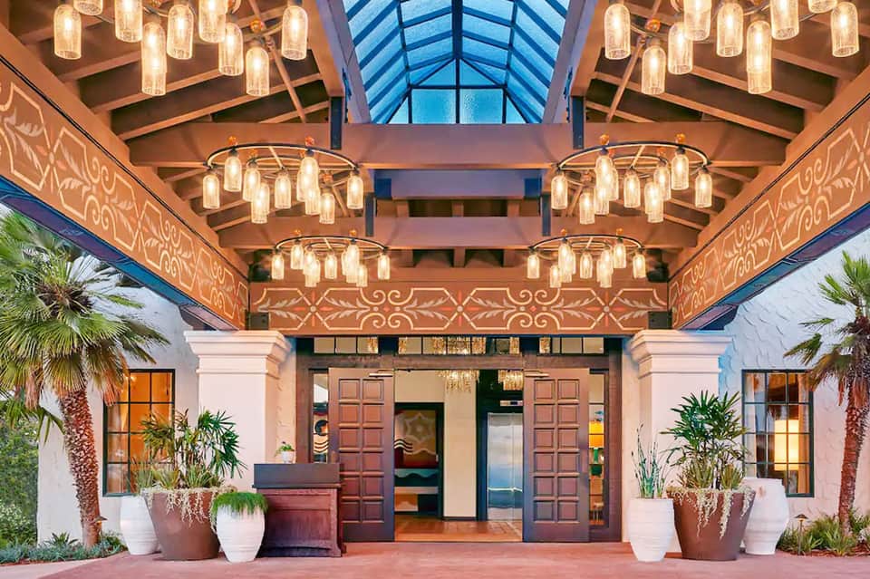 Elegant hotel entrance with ornate ceiling, brass pendant lights, palm plants, and wooden doors
