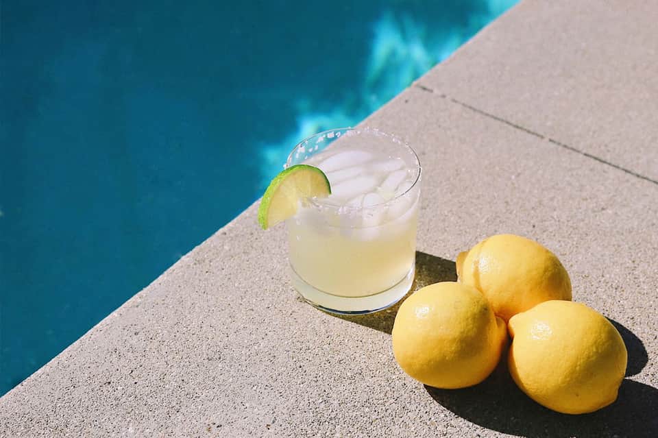 Poolside cocktail with lime slice and fresh lemons on concrete deck by blue water