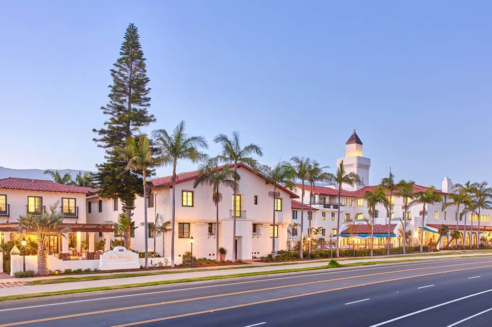 Spanish colonial-style hotel exterior with white stucco, red tile roof, palm trees, and tower at dusk