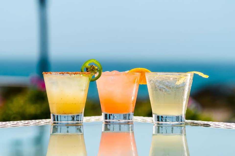 Three colorful tropical cocktails with ocean view backdrop and citrus garnishes