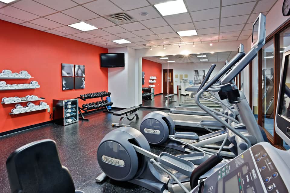 Modern gym facility with red accent walls, cardio equipment, dumbbells on racks, and workout machines