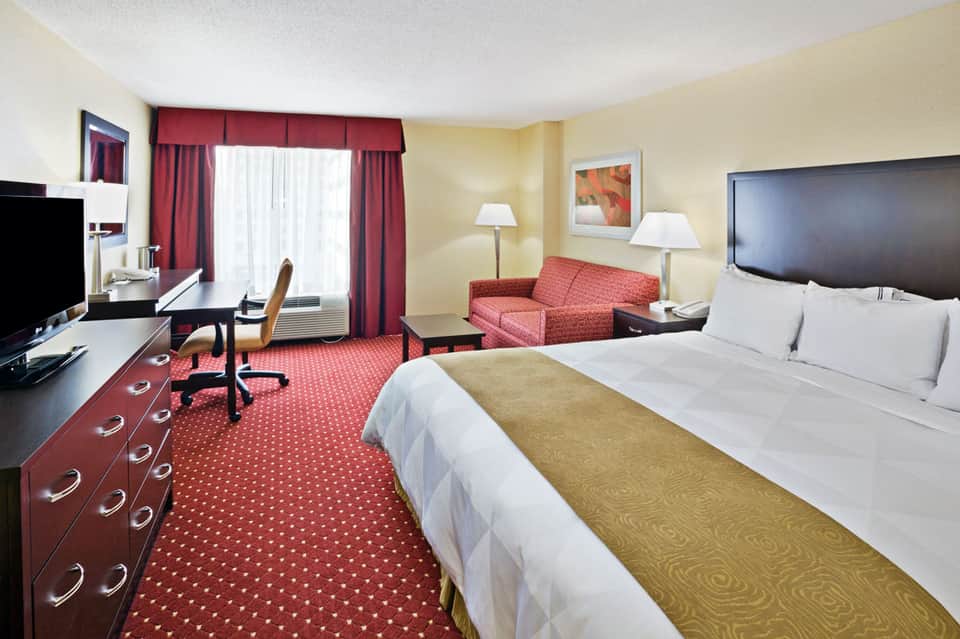 Hotel room with queen bed, red sofa, desk, flat-screen TV, and patterned red carpet