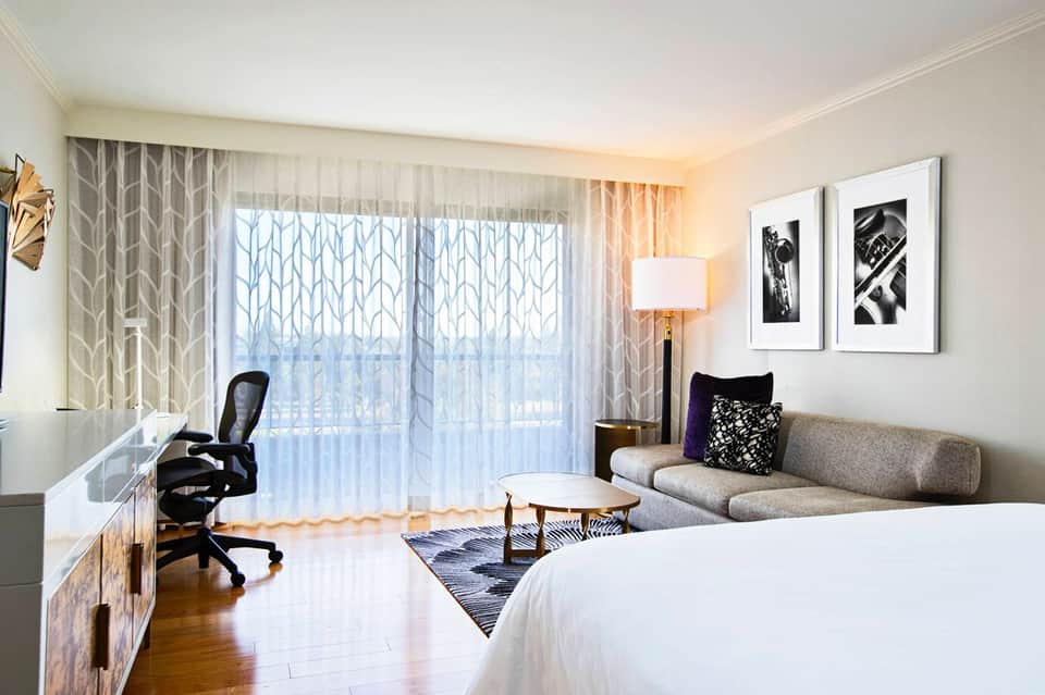 Modern hotel room with queen bed, sofa, work desk, balcony access, and contemporary furnishings
